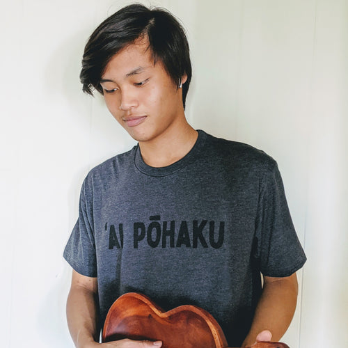 Hawaiian male  wearing a soft lightweight heather charcoal men's tee with 'AI PŌHAKU  in black ink on the chest while holding a wodden bowl